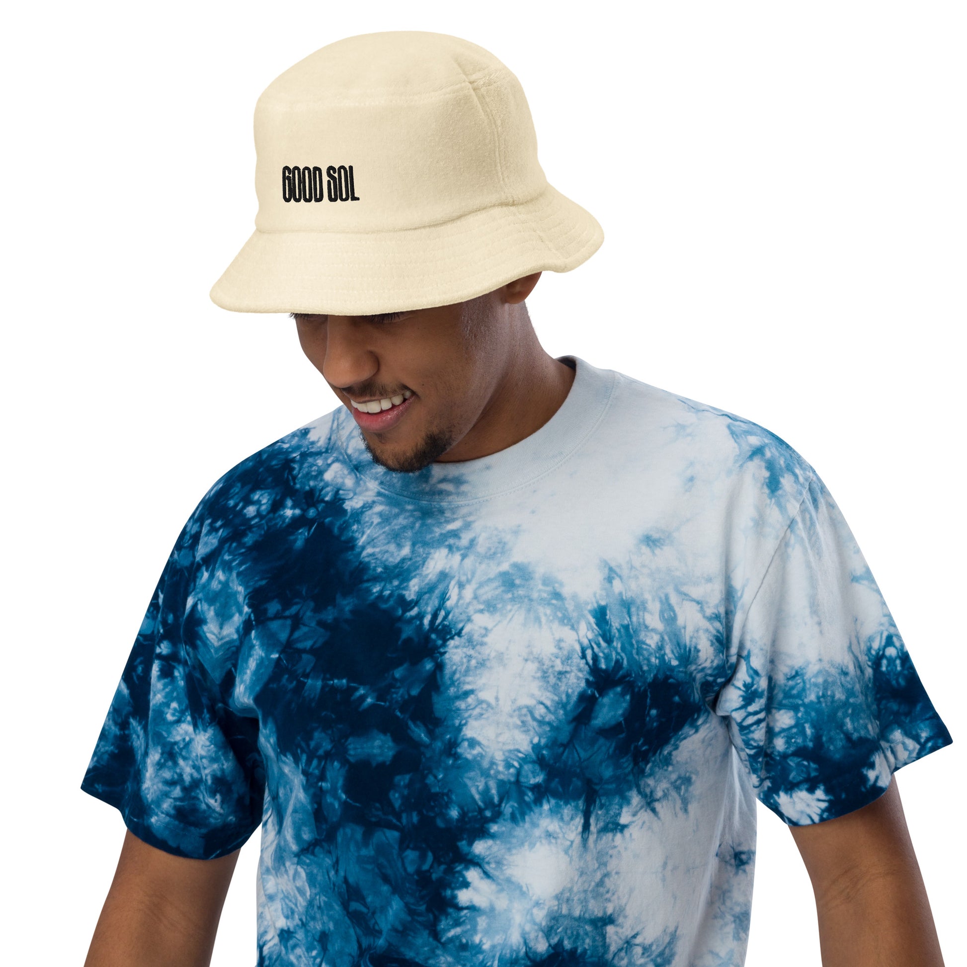 Black person with a blue tie dye shirt with a yellow terry cloth bucket hat. The word Good Sol is threaded at the top.